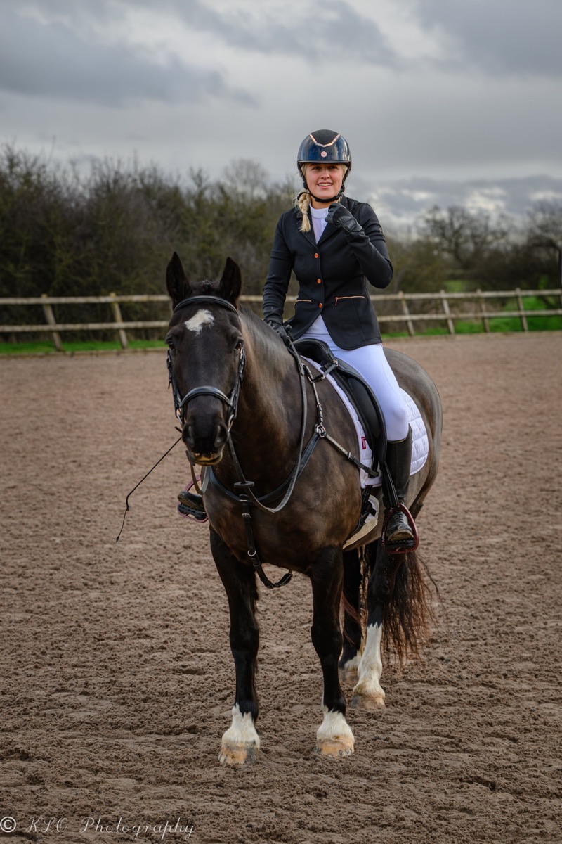 A young woman riding a black and white horse on a dirt riding arena under a cloudy sky, wearing a black riding helmet, black riding jacket, and white riding pants.