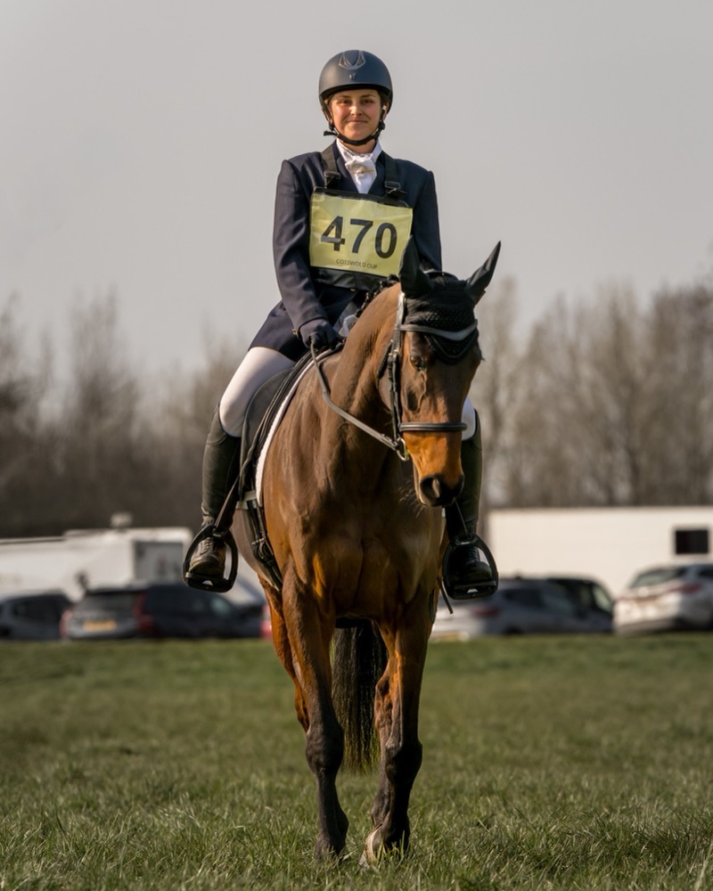 A woman riding a brown horse on a grassy field, wearing a black helmet and a navy blazer with a yellow number bib labeled 470 from the Cotswold Cup, with parked cars and trees in the background.