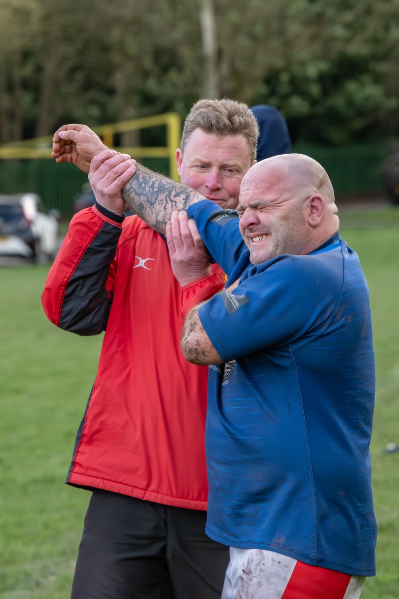 Two men are engaged in a chokehold wrestling match on a grassy field, with trees and parked cars in the background. One man is wearing a red jacket and black pants, and the other is dressed in a blue shirt and red and white shorts.