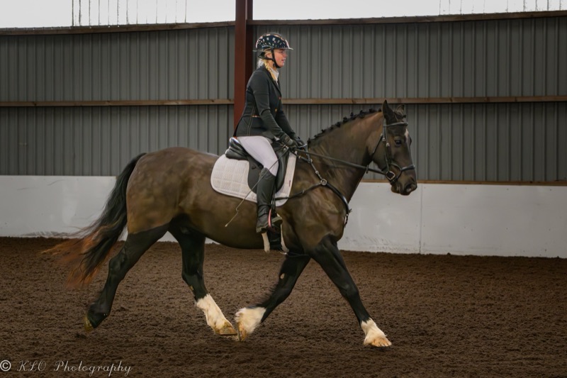 A woman riding a horse inside an indoor riding arena. The woman is wearing a riding helmet, a black blazer, white riding breeches, and black riding boots. The horse has a brown coat with a white face marking and is equipped with a saddle, bridle, and
