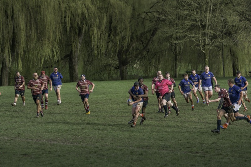 Rugby players wearing maroon and blue jerseys playing a match on a grassy field surrounded by trees.