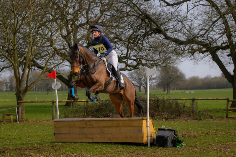 A female equestrian rider wearing a helmet and riding attire is jumping a horse over a wooden obstacle during a cross-country riding event, set outdoors with leafless trees and a grassy field in the background.