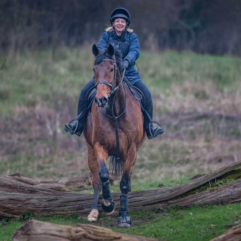 A woman riding a horse outdoors in a natural setting with trees and grass, wearing a helmet and riding gear, smiling.