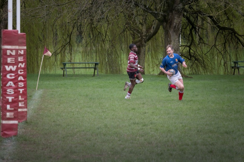 Two rugby players running towards a try zone, with a corner flag and goal post nearby, in a park with green grass and trees in the background.