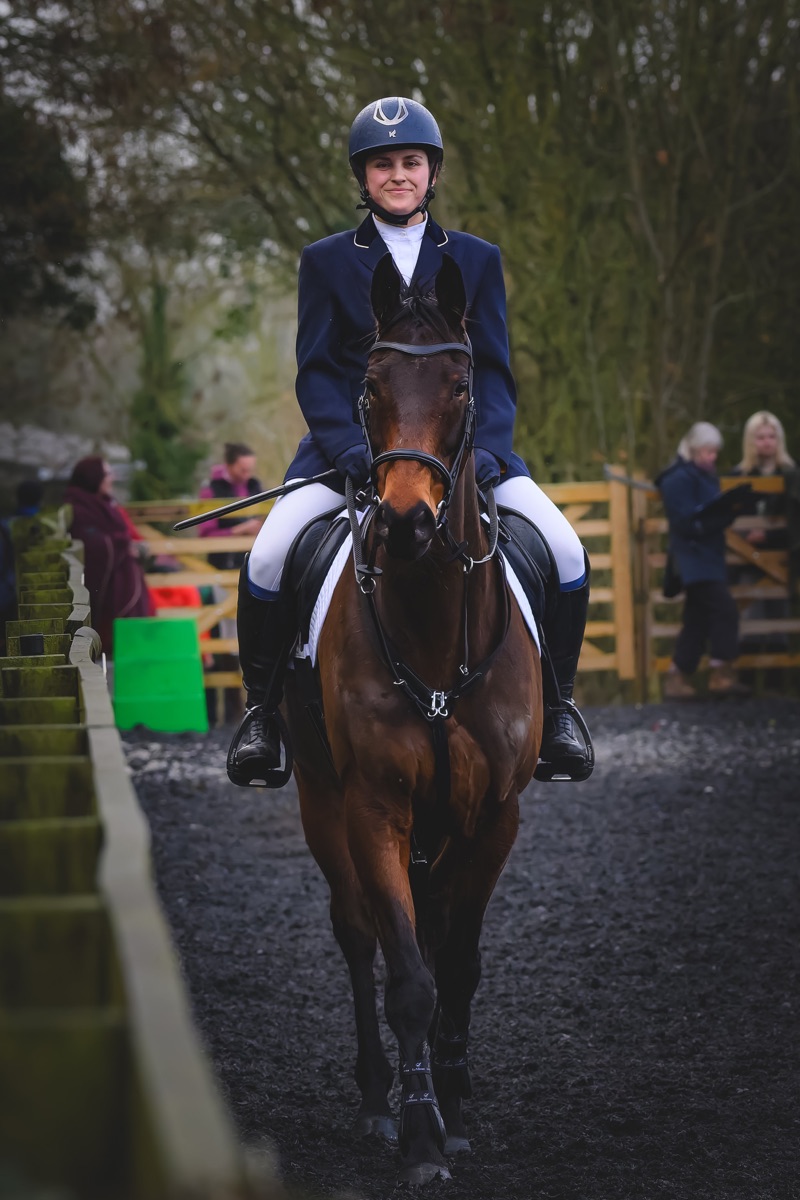 A young woman riding a horse on a trail, wearing a riding helmet and formal riding attire, with onlookers in the background.