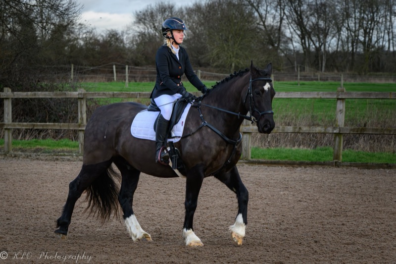 A young woman riding a black and white horse on a dirt riding arena, with a wooden fence and leafless trees in the background on a cloudy day.