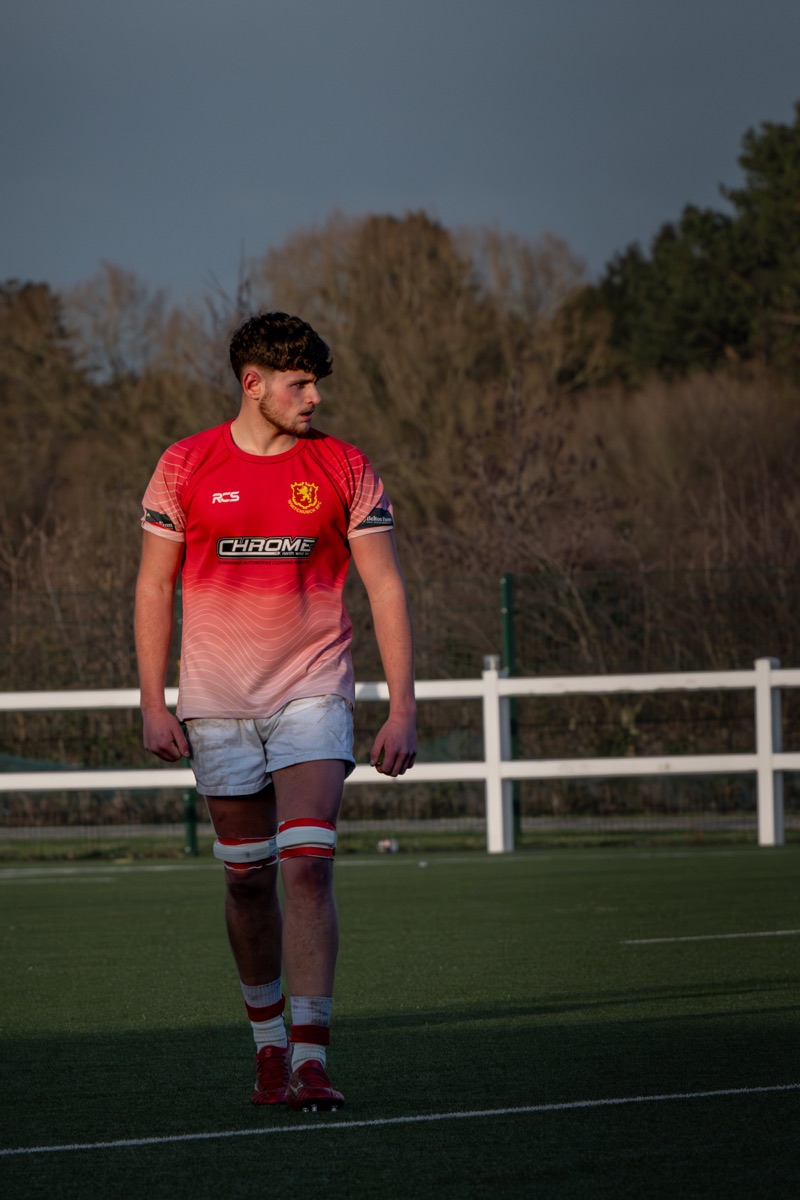 A soccer player with curly dark hair wearing a red jersey, white shorts, and red socks walking on a field with white boundary lines, a white fence, and trees in the background under a darkening sky.