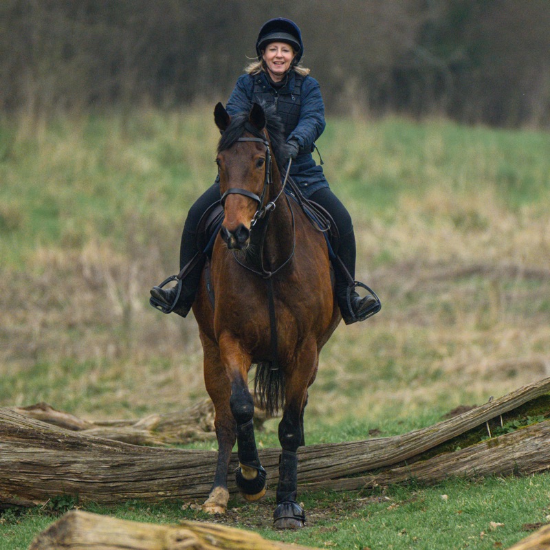 A woman riding a horse across a grassy field with a blurred background, wearing a riding helmet and navy jacket.