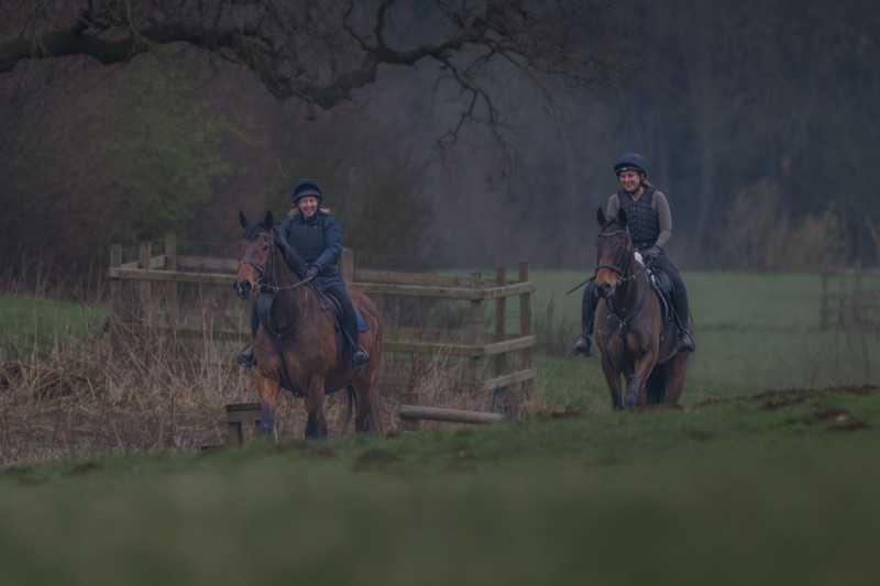 Two women riding horses in a grassy field, smiling and wearing riding helmets and vests, with a wooden fence and trees in the background.