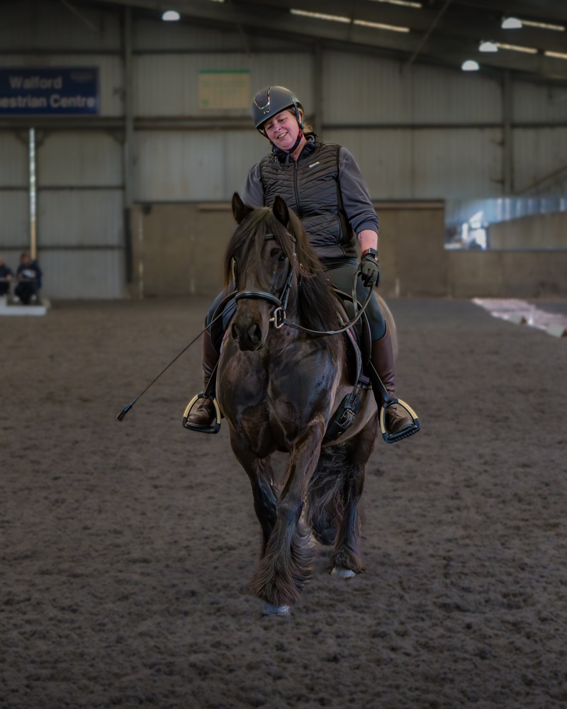 A woman riding a brown horse inside an indoor riding arena. She is wearing a helmet, a black vest, and gloves, and appears to be smiling or smiling with some discomfort.