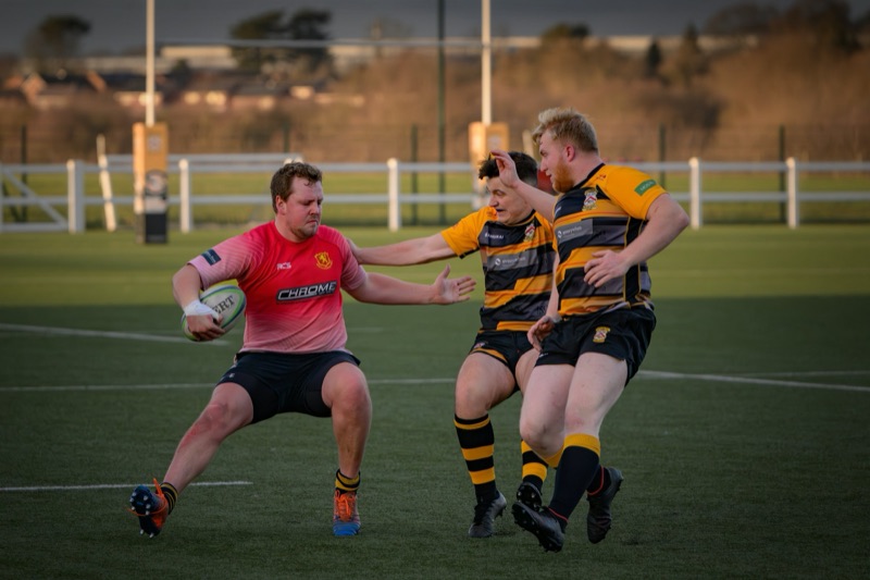 Rugby players during a match, with one player in a pink jersey holding a rugby ball and two players in black and yellow jerseys attempting to tackle him on a field.