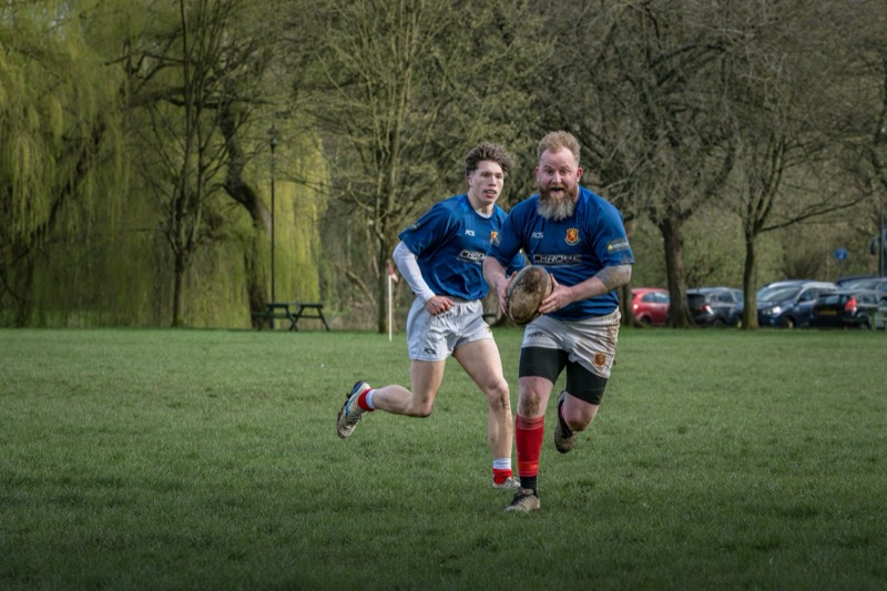 Two men playing rugby outdoors, running on a grassy field with trees and parked cars in the background. One man has a beard and is holding a rugby ball, while the other young man runs beside him.