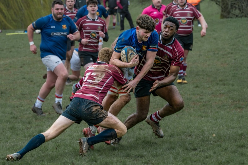 Rugby players in action during a match on a grassy field, with one player holding the ball tightly as others attempt to tackle him, surrounded by teammates.
