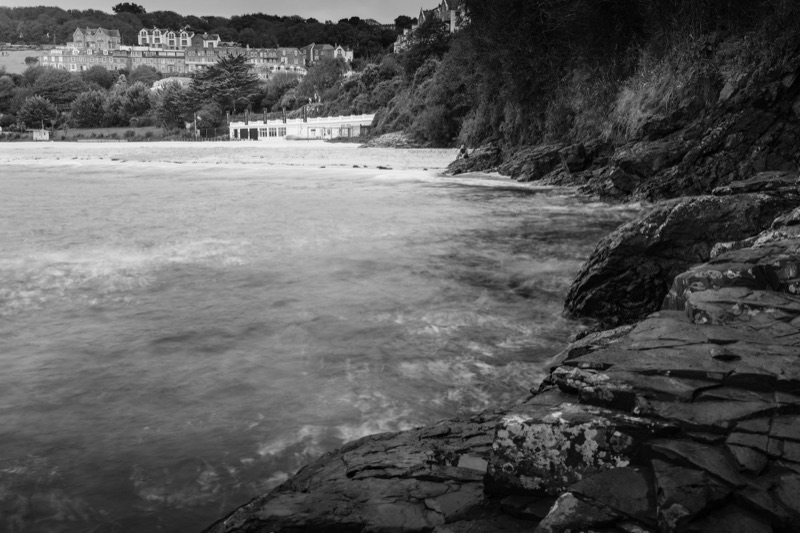 A black and white photo of a rocky coastal area with water in the foreground, a sandy beach and buildings on the far side, and a hillside with houses and trees in the background.