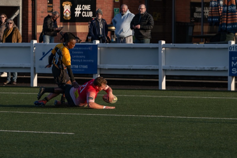A rugby player in a pink and yellow uniform on the ground holding a rugby ball, with another player in a dark uniform reaching towards her. In the background, a woman in a brown jacket and sunglasses watches, and several men stand behind a white fenc