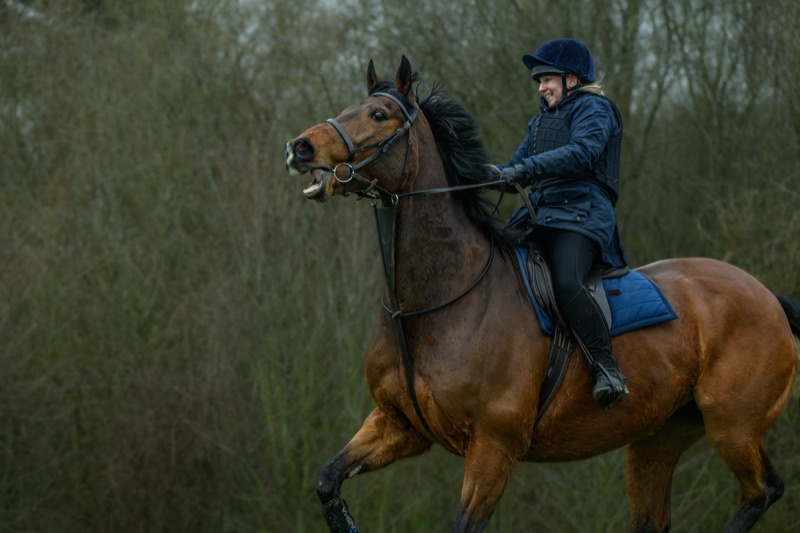A woman riding a brown horse with a dark mane through a wooded area, both appearing to enjoy the activity.