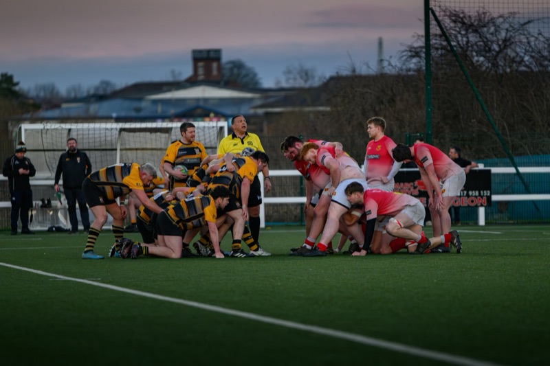 Two rugby teams, one in yellow and black jerseys and the other in pink and white jerseys, are engaging in a scrum on the field during a game at dusk. An umpire in yellow is overseeing the play, with spectators and a goal post visible in the backgroun