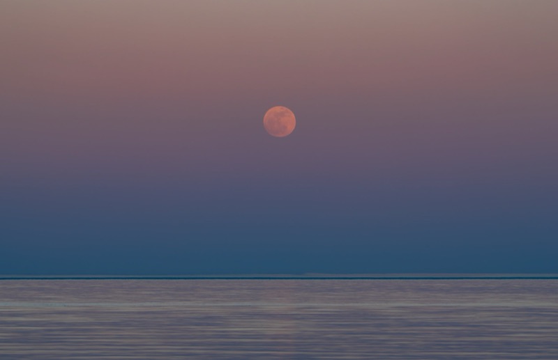 Full moon over a calm ocean at sunset with a pink and purple sky.