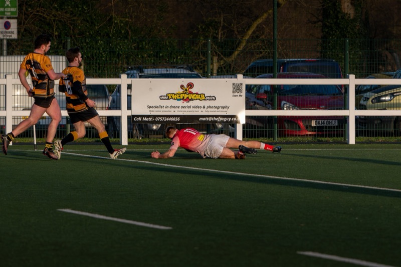 Two rugby players in yellow and black jerseys run past a fallen player in a pink jersey on a green field during a match.