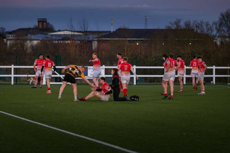 A rugby team in red and white uniforms standing and sitting on a field, with a coach or referee in a black and yellow uniform, as players tend to an injured teammate. A dog is visible in the background near the white fence, and the scene is set durin