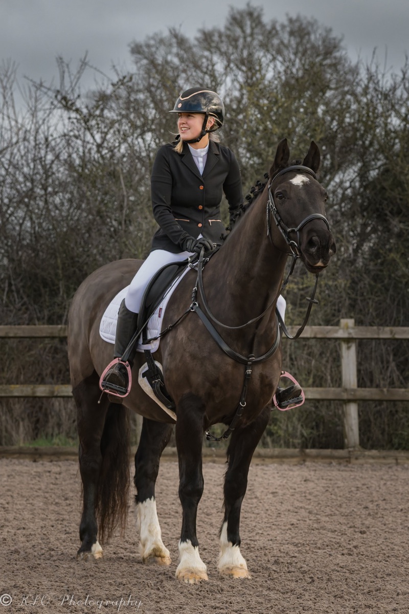 A young woman with blonde hair riding a brown and white horse in an outdoor riding arena during cloudy weather. She is wearing a black helmet, black riding jacket, white breeches, and black gloves. The horse has black and white markings, is outfitted