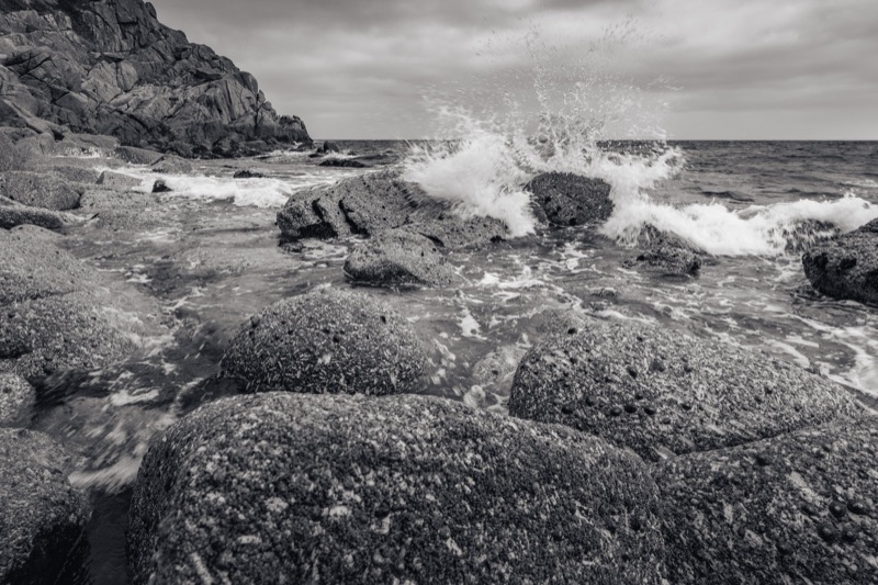 Black and white photo of a rocky seashore with waves crashing against rocks under an overcast sky.