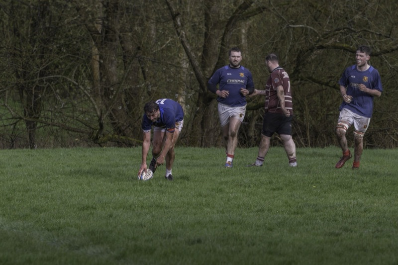 Rugby players on a muddy field, one is bent over holding a rugby ball, while three others are standing and talking, with trees in the background.