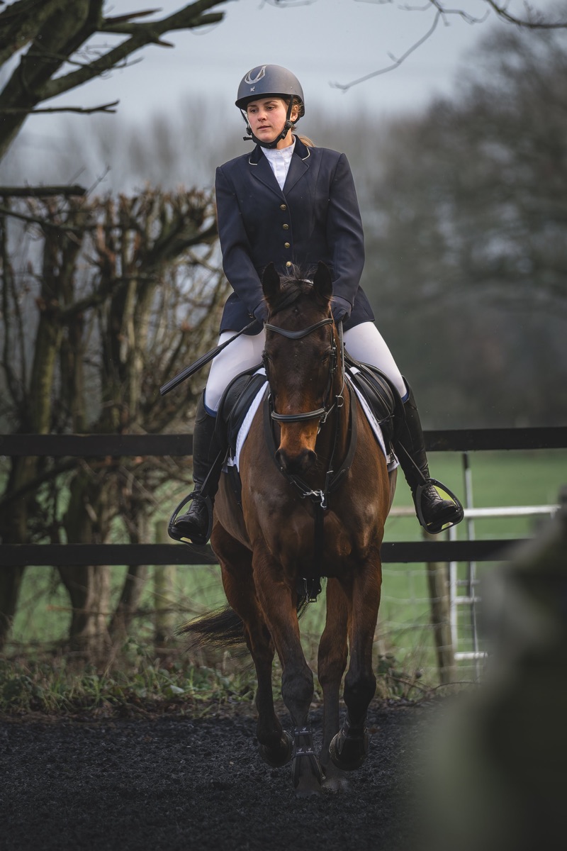 A female equestrian rider in formal riding attire on a brown horse, riding through a fenced outdoor course on a cloudy day.