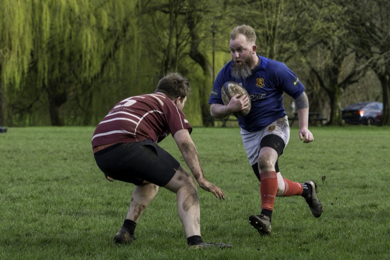 A rugby player in a blue jersey with a beard holding a rugby ball while running past a player in a maroon jersey who is crouched low on the field. The scene takes place in a park with green grass and trees in the background.