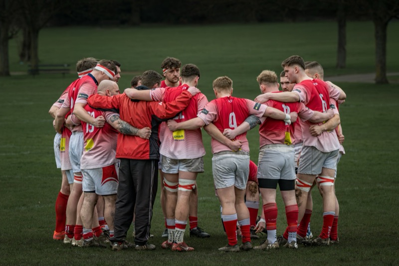 A rugby team huddle on a field during daytime, players dressed in red and white uniforms with some wearing headbands, standing close together with arms around each other.
