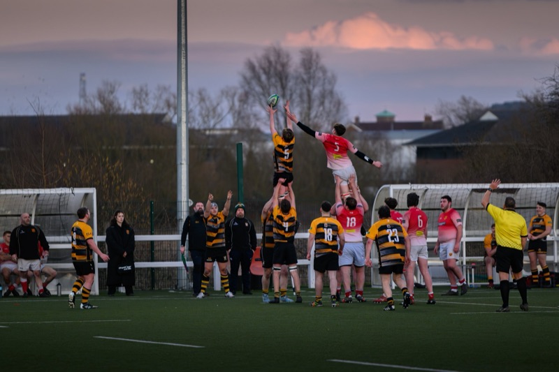 A rugby match with players jumping to catch the ball, some players are in yellow and black jerseys, while others have pink jerseys. The game is taking place on a field with a crowd and coaches watching, under a partly cloudy sky at sunset.