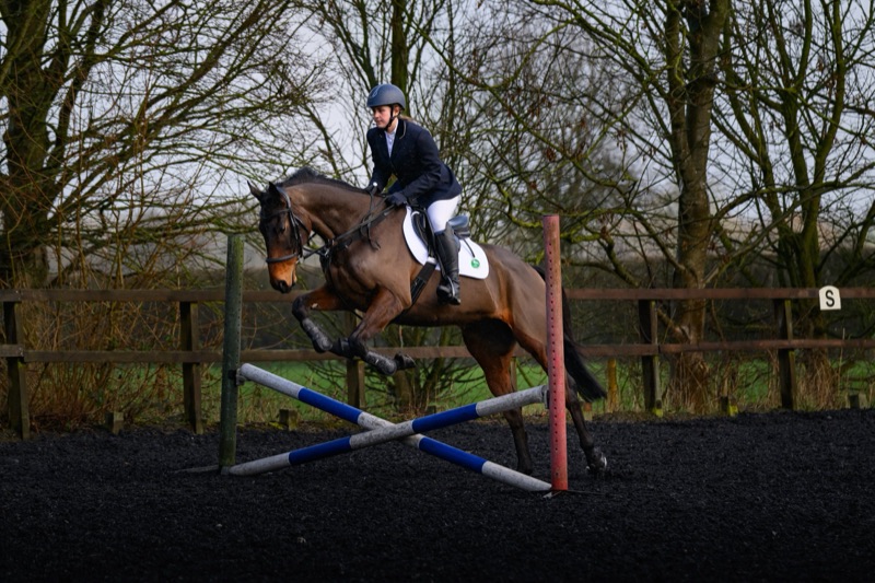 A woman wearing formal riding attire, including a helmet, riding a brown horse over a showjumping obstacle outdoors during daytime.