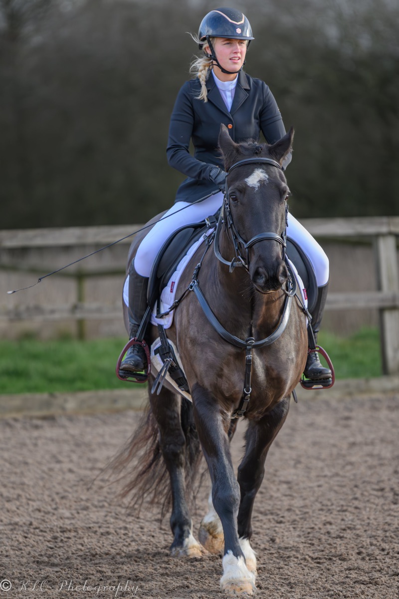 A young woman riding a dark-colored horse with a white star on its forehead, wearing a helmet and riding gear, in an outdoor riding arena.