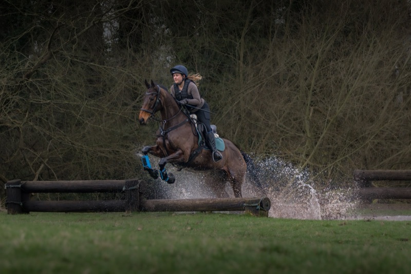 A woman riding a horse jumps over a wooden obstacle in an outdoor setting surrounded by trees.
