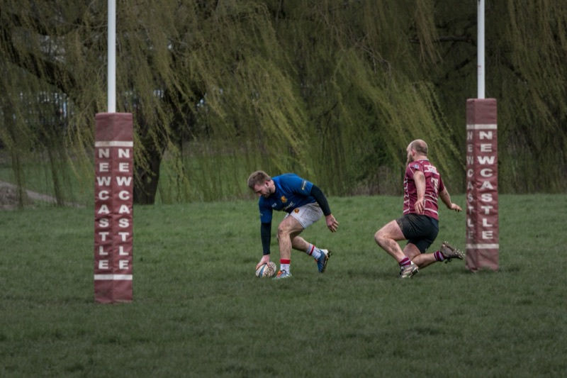 Two rugby players on a grassy field, one in a blue jersey and the other in a maroon jersey, near the rugby goal post, during daylight.