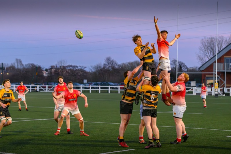 Rugby players in action on a field during a match, with players jumping to catch the ball as others support from below.