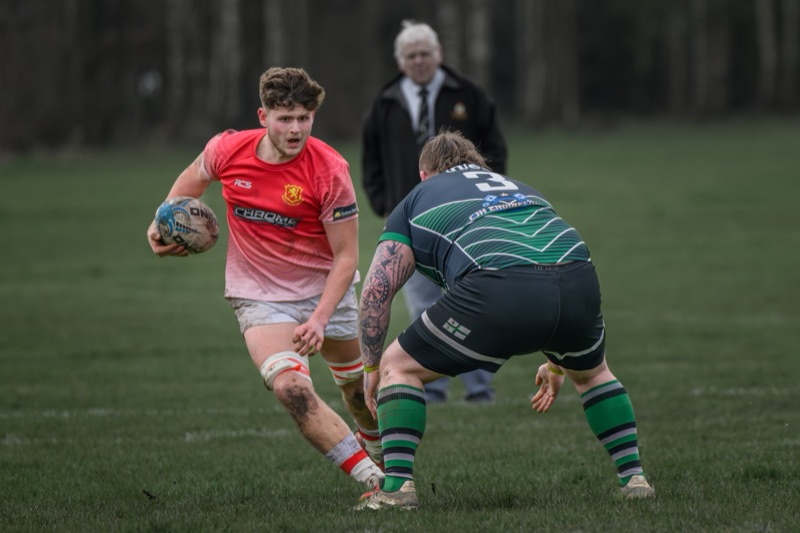 A rugby match in progress with a player in a red jersey holding a rugby ball, running forward while an opponent in a dark jersey with green and white striped socks is attempting to tackle him. An official watches in the background on a grassy field w