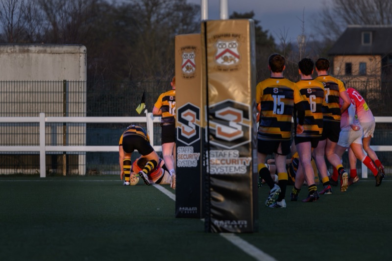A rugby match in progress with players on the field, some kneeling and others standing, near the goal post at dusk. One player is on the ground holding the ball close to the try line while others are gathered or walking nearby.