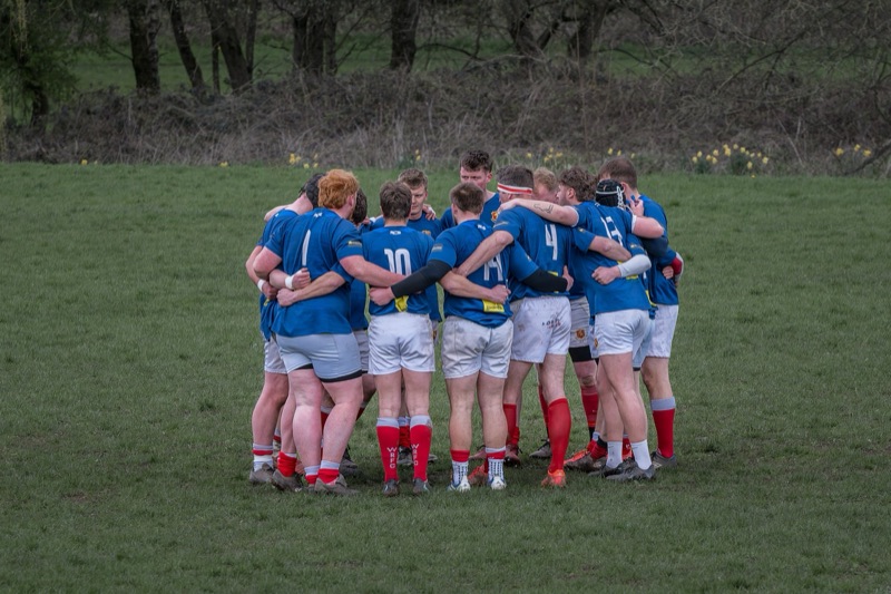A group of rugby players wearing blue jerseys and white shorts huddling together on a grassy field, with trees in the background.