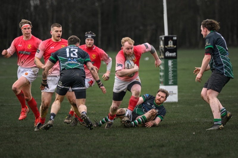 Rugby players in action during a match, with one player holding a rugby ball and others tackling or running on a grassy field.