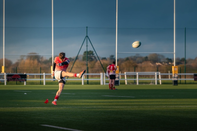 A rugby player in a red jersey and white shorts kicking a rugby ball during a game on a field.
